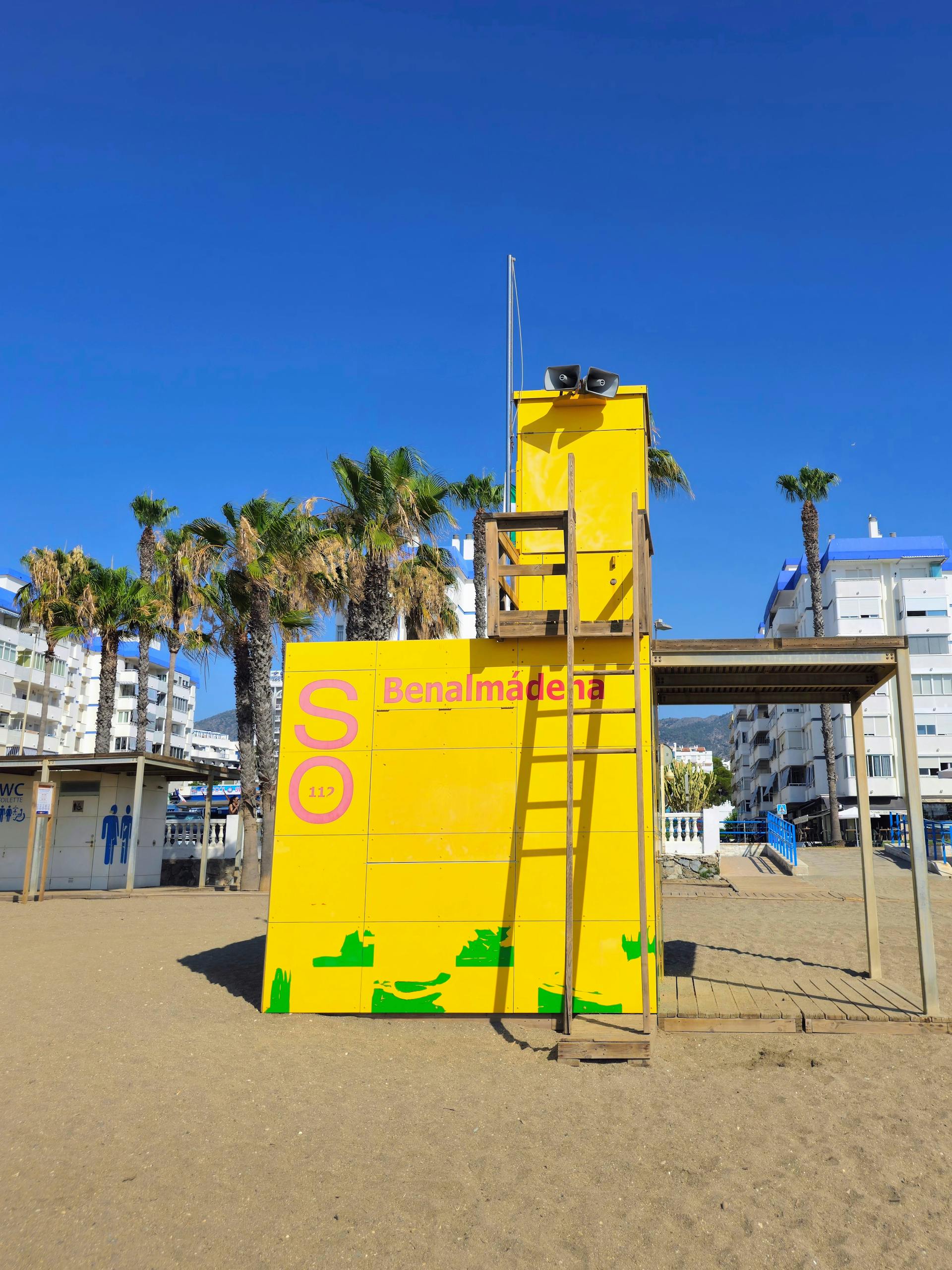 Stafford Affiliates Travel - A bright yellow lifeguard tower on a sandy beach in Benalmádena, Spain, under a clear blue sky.