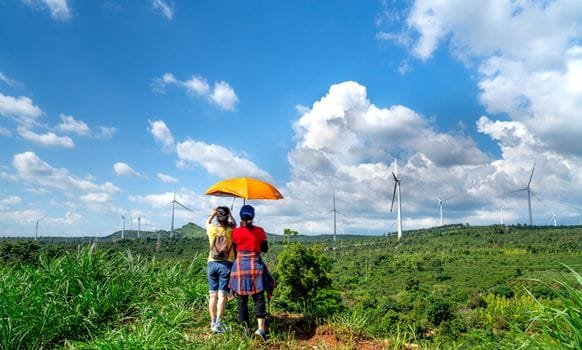 Two travellers standing on a lush green hill under an orange umbrella, observing wind turbines on a sustainable eco-tourism landscape – Stafford Affiliates Travel.