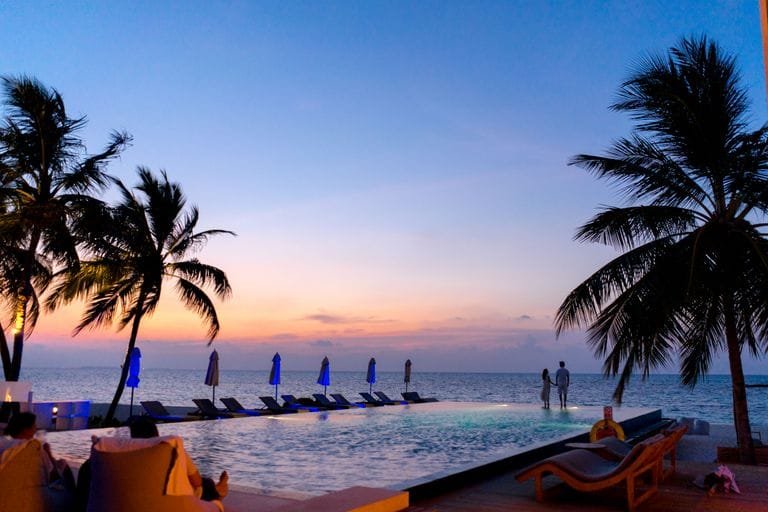 A couple enjoys a picturesque sunset by the seaside infinity pool, surrounded by palm trees. - broadwaytravel.com - Stafford Affiliates Travel