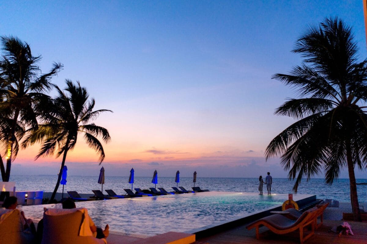 A couple enjoys a picturesque sunset by the seaside infinity pool, surrounded by palm trees. - broadwaytravel.com - Stafford Affiliates Travel