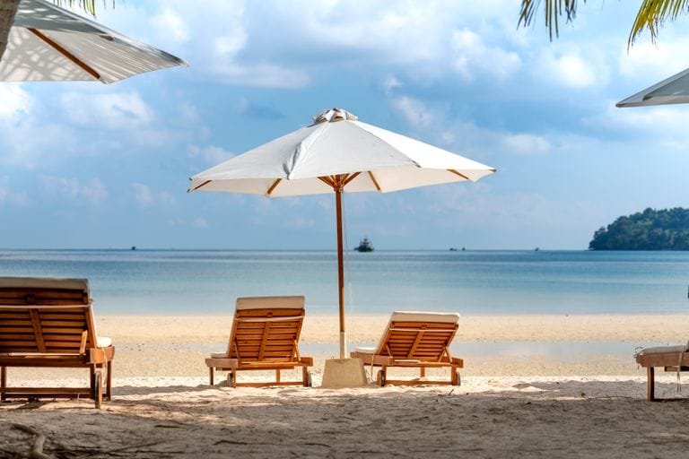Amazing scenery of tropical sandy beach with cozy wooden sunbeds under umbrellas against picturesque blue sea on clear sunny day- Destination2 Review: UK Holiday Booking Guide / Stafford Affiliates Travel