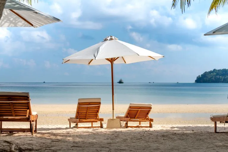 Amazing scenery of tropical sandy beach with cozy wooden sunbeds under umbrellas against picturesque blue sea on clear sunny day- Destination2 Review: UK Holiday Booking Guide / Stafford Affiliates Travel